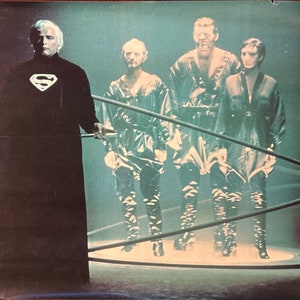 Puede incluir: Un hombre con un traje negro con el logotipo de Superman en el pecho se encuentra frente a otras tres personas vestidas de negro. El grupo está rodeado por una estructura circular brillante.