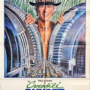 Może przedstawiać: Plakat filmowy do filmu "Krokodyl Dundee" przedstawiający Paula Hogana w kapeluszu bushman, patrzącego z dżungli na ruchliwą ulicę w Nowym Jorku. Tekst na plakacie brzmi: "Paul Hogan to Krokodyl Dundee. Trochę go jest w każdym z nas."