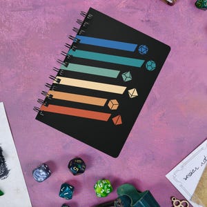 May include: A black spiral notebook with a rainbow stripe design and dice icons. The notebook is on a pink surface with dice scattered around it.