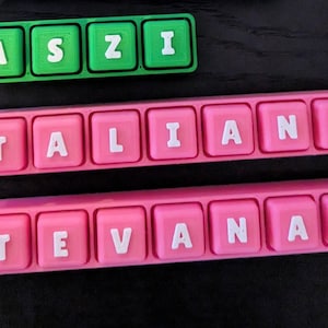 May include: Three personalized name keychains. One green keychain spells out "ASZI" in white letters. Two pink keychains spell out "TALIANA" and "TEVANAIA" in white letters. Each keychain has a matching colored chain.