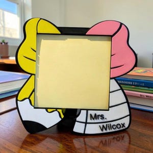 May include: A square photo frame featuring a stylized pencil design with a yellow top, a pink eraser section, and a white body. The frame includes black outlines and the text Mrs. Wilcox printed in black on the lower right side of the white pencil base.