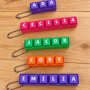 May include: Five rectangular keychains featuring colorful plastic blocks with white letters spelling the names ADA, CECILIA, JACOB, CORY, and EMILIA. Each keychain has a silver metal ball chain attached to one end for hanging purposes.