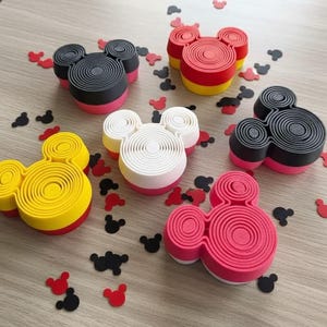 May include: Six three-dimensional mouse-shaped containers in red, yellow, black, white, and pink, each featuring a concentric circular pattern on the top surface. Small mouse-shaped confetti pieces in red and black are scattered around the containers.
