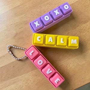 May include: Three rectangular silicone keychains with raised white letters. One purple keychain displays XOXO, one yellow keychain displays CALM, and one pink keychain displays LOVE. The pink keychain includes a silver-toned metal ball chain attachment.