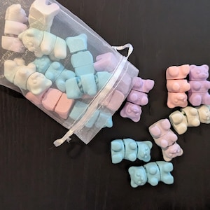 May include: A collection of pastel-colored gummy bear candies in a small, white mesh bag. The bears are pink, blue, purple, and white. Some are stacked in small piles on a dark surface.