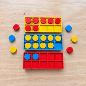 May include: Three rectangular ten-frame trays in yellow, blue, and red, each containing ten square compartments. The yellow tray holds seven red circular counters, the blue tray holds nine yellow circular counters, and the red tray holds two blue circular counters. Additional loose counters are scattered around the trays.