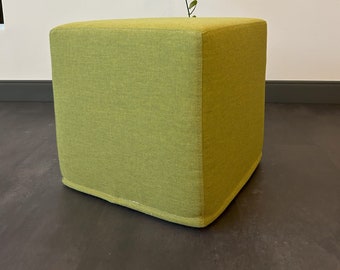 May include: A square green fabric ottoman with a subtle texture. The ottoman is sitting on a dark gray floor.