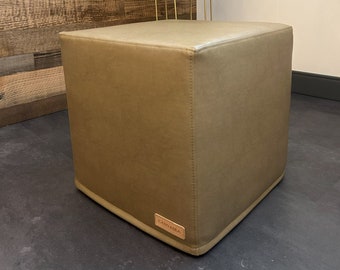 May include: A square, light brown, upholstered ottoman with a brown leather label that says "CARVASA" on the front. The ottoman is sitting on a dark gray floor.