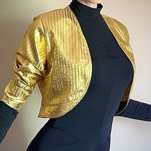 May include: A gold metallic cropped jacket with a striped pattern. The jacket has long sleeves and a slightly open front.