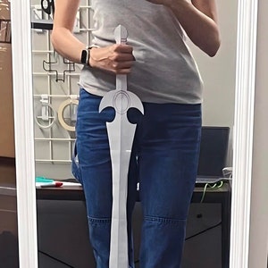 Puede incluir: Una espada de papel blanco con un mango decorativo y una punta puntiaguda. La espada está siendo sostenida por una persona que lleva un top gris sin mangas y pantalones vaqueros azules.