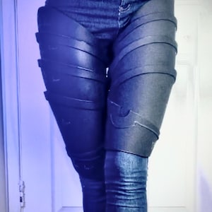 Könnte beinhalten: Dunkle Jeans mit schwarzen, rüstungsartigen Beinbedeckungen. Die Beinbedeckungen haben ein geschichtetes Design mit dekorativen Akzenten. Die Rüstung scheint aus einem starren Material zu bestehen und ist mit Riemen befestigt.