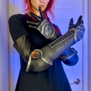 May include: A pair of black and silver cosplay arm gauntlets with a detailed design. The gauntlets have a textured surface and a silver trim. The gauntlets are worn on both arms and have a mechanical look.