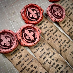 May include: Close-up of several pink wax seals with a fleur-de-lis design, alongside fabric strips printed with text. The seals have a distressed, aged appearance. The text on the fabric reads "Emperor, bestow on me..." in a gothic font.