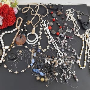 May include: A selection of necklaces in diverse designs and materials, such as pearls, beads, and wooden pendants. The necklaces are displayed on a dark surface, with a bouquet of red and pink flowers in the background.