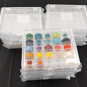 Könnte beinhalten: Transparenter Kunststoff-Nähgarn-Organizer mit einer Vielzahl farbiger Garnspulen. Der Organizer ist rechteckig mit einem Klappdeckel und in einer transparenten Plastiktüte verpackt. Mehrere zusätzliche Organizer sind in der Nähe gestapelt.