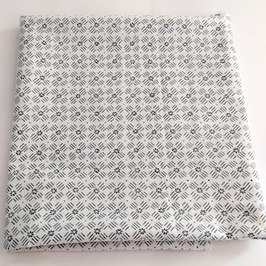 May include: Folded white fabric featuring a repeating pattern of small black floral and geometric designs. The fabric is neatly folded, displaying the detailed pattern. The overall aesthetic is clean and minimalist.