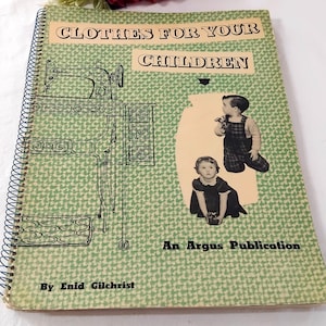 May include: A vintage spiral-bound book titled "Clothes For Your Children" with a green patterned cover. The cover features illustrations of a sewing machine and children. The book is an Argus Publication, written by Enid Gilchrist.