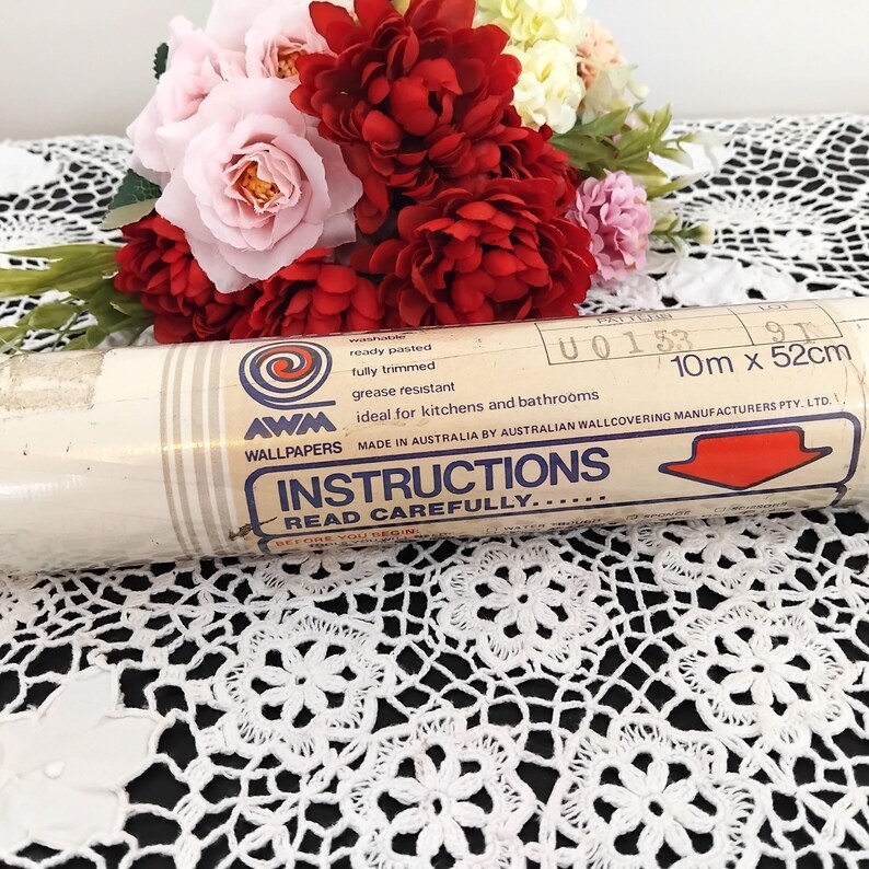 May include: A roll of AWM Wallpapers with instructions, 10m x 52cm, ideal for kitchens and bathrooms. The label is cream-colored with blue and red text. A bouquet of red and pink flowers sits behind the wallpaper roll.