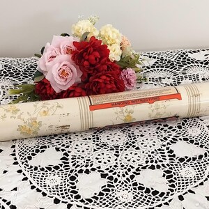 May include: A rolled-up vintage wallpaper roll with a cream background and yellow floral pattern. The roll has a label with "Hanging Instructions" printed on it. A bouquet of pink and red flowers sits nearby on a white lace tablecloth.