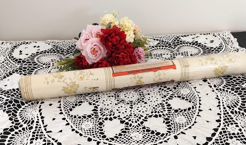 May include: A rolled-up vintage wallpaper roll with a cream background and yellow floral pattern. The roll has a label with "Hanging Instructions" printed on it. A bouquet of pink and red flowers sits nearby on a white lace tablecloth.