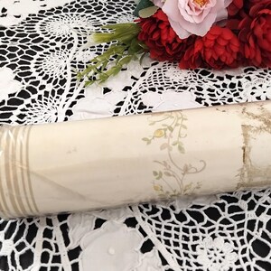 May include: A rolled-up vintage wallpaper roll. The wallpaper is cream-colored with a floral pattern in yellow and green. The roll is wrapped in clear plastic and has vertical beige stripes on one end.