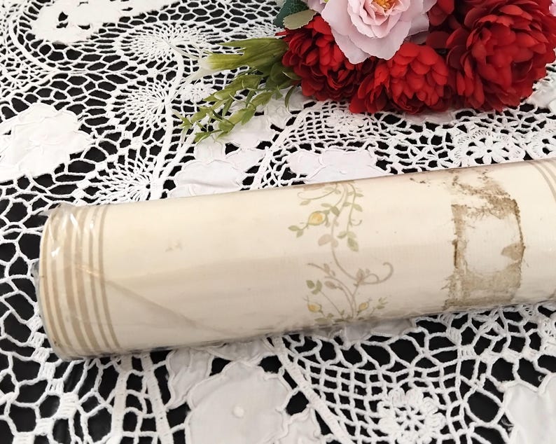 May include: A rolled-up vintage wallpaper roll. The wallpaper is cream-colored with a floral pattern in yellow and green. The roll is wrapped in clear plastic and has vertical beige stripes on one end.