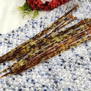 May include: A set of knitting needles with a tortoiseshell design. The needles have a pointed end and a small, rounded orange tip. They are placed on a white and blue textured fabric.