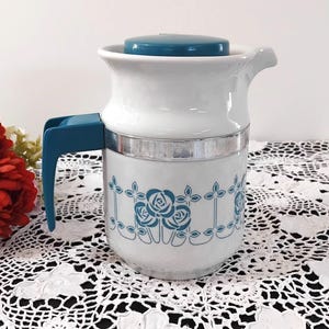 May include: White ceramic jug with a teal lid and handle. The jug features a blue and white floral design with a silver band around the middle.