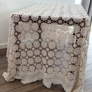 May include: A cream-coloured, crocheted tablecloth draped over a wooden table. The tablecloth has a repeating floral pattern and a scalloped edge. The intricate design adds a touch of vintage charm to the table setting.