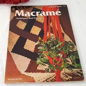 May include: A softcover book titled Macramé Techniques and Projects, A Sunset Book. The cover features a photograph of a bright orange macramé plant hanger holding a green plant, set against a background of a brown, black, and cream woven rug.