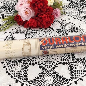 May include: A roll of vintage Duralon vinyl wallcovering with a floral pattern. The label reads "NEW" and "vinyl wallcovering". The roll is cream-colored with a floral design and is resting on a white lace tablecloth. A bouquet of red and pink flowers is in the background.