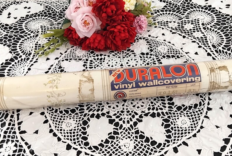 May include: A roll of vintage Duralon vinyl wallcovering with a floral pattern. The label reads "NEW" and "vinyl wallcovering". The roll is cream-colored with a floral design and is resting on a white lace tablecloth. A bouquet of red and pink flowers is in the background.