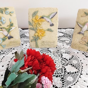 May include: Three rectangular plaques depicting hummingbirds and flowers. Each plaque shows a different hummingbird and floral arrangement in relief against a textured background. The plaques are displayed on clear stands, with a bouquet of red flowers in the foreground.