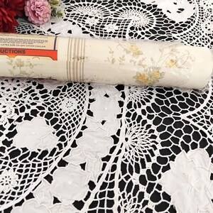 May include: A roll of wallpaper with a floral pattern featuring yellow roses and green leaves. The wallpaper has a cream background with vertical beige stripes. The roll is partially wrapped in plastic and rests on a white lace tablecloth.