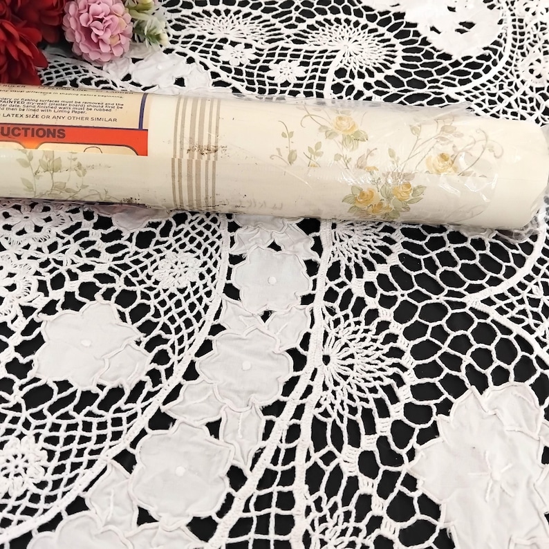 May include: A roll of wallpaper with a floral pattern featuring yellow roses and green leaves. The wallpaper has a cream background with vertical beige stripes. The roll is partially wrapped in plastic and rests on a white lace tablecloth.