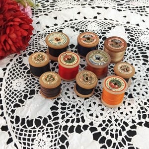 May include: A collection of eleven vintage wooden spools of sewing thread in various colours including black, red, orange, and tan. Each spool features a circular paper label on top with printed brand text such as J&P Coats, Sylko, and Phoenix.