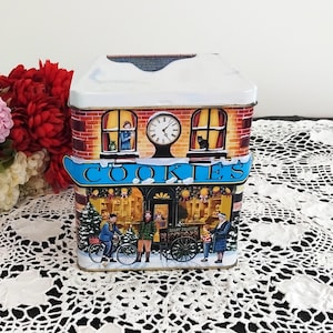 May include: A rectangular cookie tin depicting a winter scene. The tin is shaped like a building with a snow-covered roof, brick facade, and a clock. The word "COOKIES" is displayed on a blue sign. The scene depicts people and a Christmas tree.