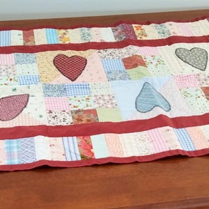 May include: A rectangular patchwork table runner featuring a variety of small floral, plaid, and gingham fabric squares in pastel and muted tones. The runner is bordered by a solid dark red fabric and decorated with six appliqued heart shapes.