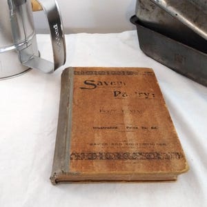May include: A vintage hardback book titled Savoury Pastry by Fred T. Vine with a tan cover, dark brown decorative borders, and black text. The book is positioned next to a silver metal flour sifter and two stacked dark metal rectangular baking tins.