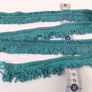 Peut inclure: Garniture de franges turquoise avec un bord supérieur tissé et des franges pendantes. Un mètre ruban indique la longueur de la garniture. La garniture est d'une couleur turquoise vibrante, et les franges sont espacées uniformément.