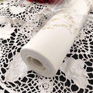 May include: A roll of off-white wallpaper with a delicate floral pattern in muted green and gold tones. The wallpaper is wrapped in clear plastic, showcasing the design. The roll is resting on a white lace tablecloth with a black background.