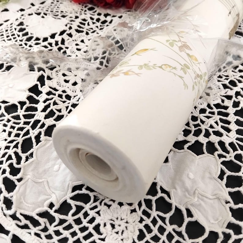 May include: A roll of off-white wallpaper with a delicate floral pattern in muted green and gold tones. The wallpaper is wrapped in clear plastic, showcasing the design. The roll is resting on a white lace tablecloth with a black background.