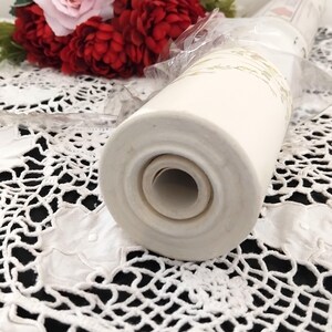 May include: A cream-colored, cylindrical ceramic object with a floral design, possibly a vase or decorative item. The object is partially wrapped in clear plastic and sits on a white lace surface. Red and pink flowers are in the background.