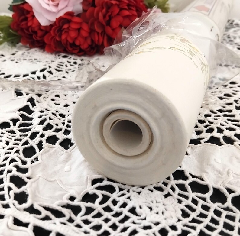 May include: A cream-colored, cylindrical ceramic object with a floral design, possibly a vase or decorative item. The object is partially wrapped in clear plastic and sits on a white lace surface. Red and pink flowers are in the background.