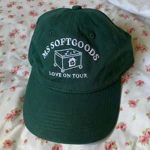 May include: A dark green baseball cap with white embroidery that reads "Ms Softgoods Love On Tour". The embroidery also includes a small illustration of a house in a box on wheels.