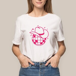 Pu&ograve; includere: Una maglietta bianca con una grafica rosa di una palla da discoteca con un cappello da cowboy in cima. La grafica &egrave; circondata da piccole stelle rosa.