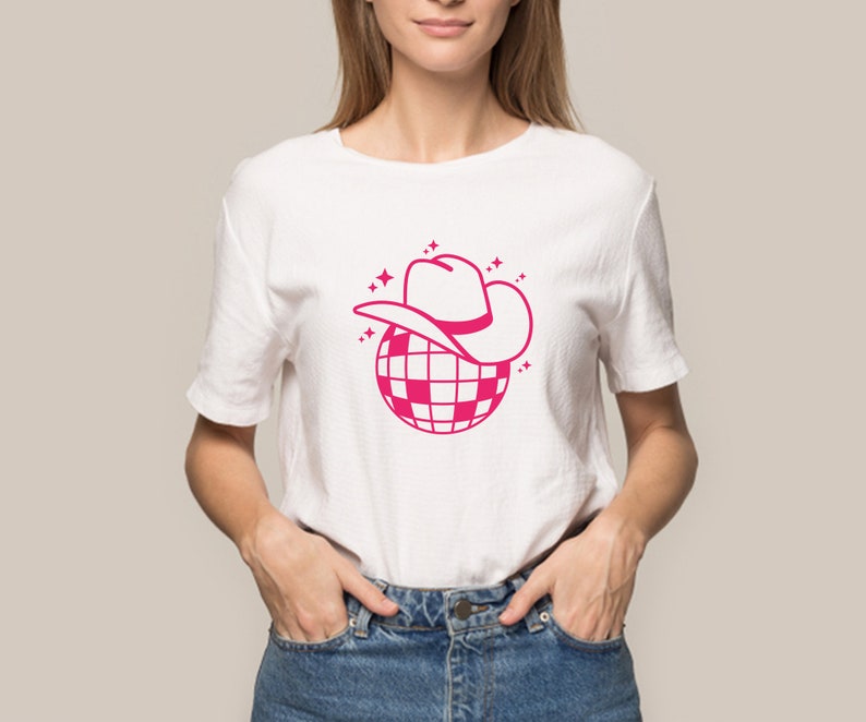 Pu&ograve; includere: Una maglietta bianca con una grafica rosa di una palla da discoteca con un cappello da cowboy in cima. La grafica &egrave; circondata da piccole stelle rosa.