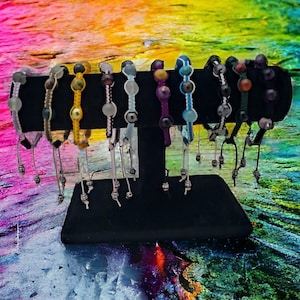 May include: A display of colorful beaded bracelets with various colors and patterns. The bracelets are made with different materials, including beads, string, and metal. The bracelets are displayed on a black stand against a colorful background.