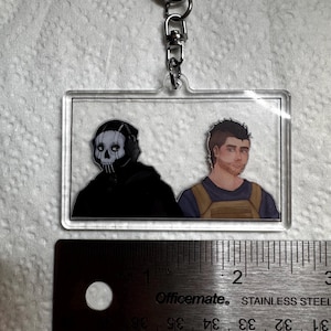 May include: A rectangular clear acrylic keychain featuring two illustrated figures. One figure is in a black hooded outfit with a skull mask, and the other is a man in a blue shirt and tan vest. The keychain is attached to a silver metal clasp.