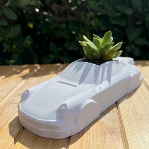 Puede incluir: Maceta de cerámica blanca con forma de coche con una pequeña planta suculenta creciendo en su interior. La maceta está sobre una superficie de madera.